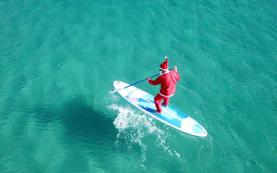 Als Weihnachtsmann verkleideter Mann paddelt auf einem Surfbrett im türkisblauen Meer in Australien.